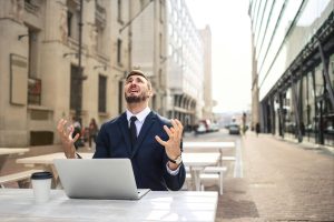 Businessman in suit showing frustration at outdoor table with laptop and coffee.